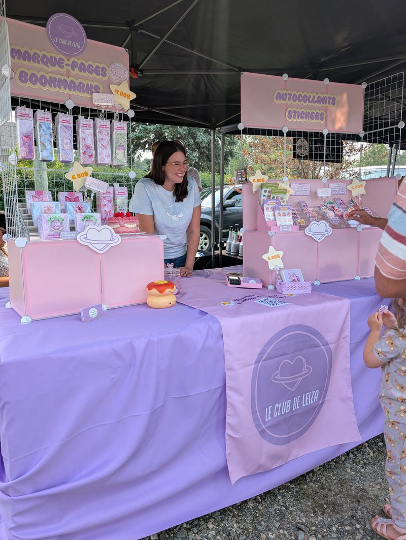 Woman standing behind a pink and purple table with products and signs at an outdoor event.